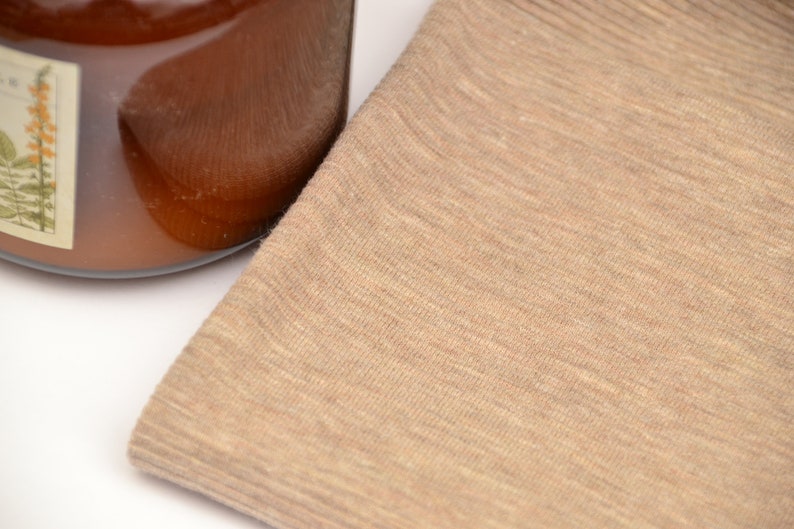 Puede incluir: Una pieza de tela de color tostado claro doblada con una textura fina y jaspeada. Junto a la tela hay una parte de un recipiente de vidrio lleno de un l&iacute;quido &aacute;mbar oscuro, con una etiqueta rectangular que muestra una ilustraci&oacute;n bot&aacute;nica de flores amarillas.