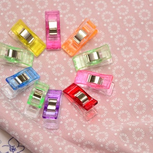 Könnte beinhalten: Eine Ansammlung farbiger Nähclips, kreisförmig angeordnet. Die Clips sind in verschiedenen transparenten Farben gehalten, darunter Gelb, Rosa, Orange, Grün, Blau, Lila und Rot, jeweils mit einem silbernen Metallverschluss. Sie liegen auf einem rosa Stoff mit weißem Blumenmuster.
