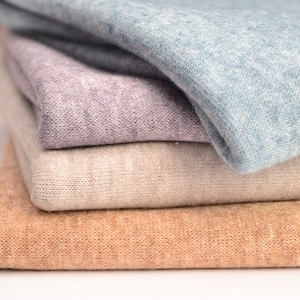 Könnte beinhalten: Ein Stapel aus vier gefalteten Stoffstücken mit weicher Strickstruktur. Die Farben von oben nach unten sind Hellblau, sanftes Lavendel, helles Beige und warmes Hellbraun. Jedes Stoffstück ist ordentlich geschichtet und zeigt ein feines Strickmuster.