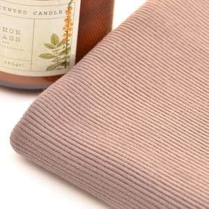 Puede incluir: Una pieza doblada de tela acanalada color rosa polvoriento con una textura vertical fina. Junto a la tela hay un frasco de vidrio marr&oacute;n de vela arom&aacute;tica con una etiqueta que dice Scented Candle Lemon Grass y una ilustraci&oacute;n bot&aacute;nica de flor amarilla.
