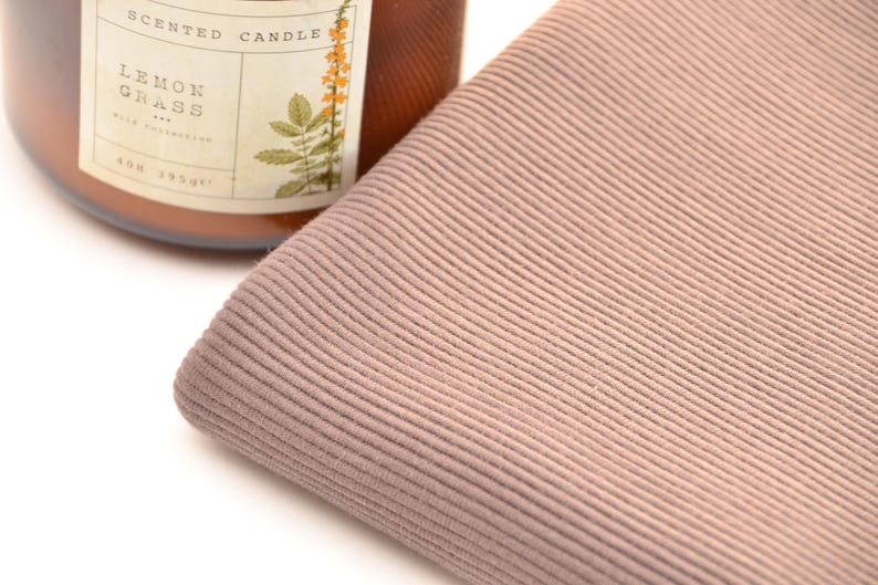Puede incluir: Una pieza doblada de tela acanalada color rosa polvoriento con una textura vertical fina. Junto a la tela hay un frasco de vidrio marr&oacute;n de vela arom&aacute;tica con una etiqueta que dice Scented Candle Lemon Grass y una ilustraci&oacute;n bot&aacute;nica de flor amarilla.