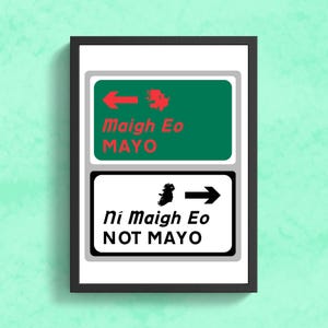 May include: A white framed print with two green and white signs. The top sign has a red arrow pointing left and the text "Maigh Eo MAYO". The bottom sign has a black arrow pointing right and the text "Ni Maigh Eo NOT MAYO".