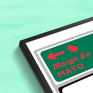 May include: A framed print of a green and white road sign with a red arrow pointing left. The sign reads "maigh Eo MAYO" in red and white text.
