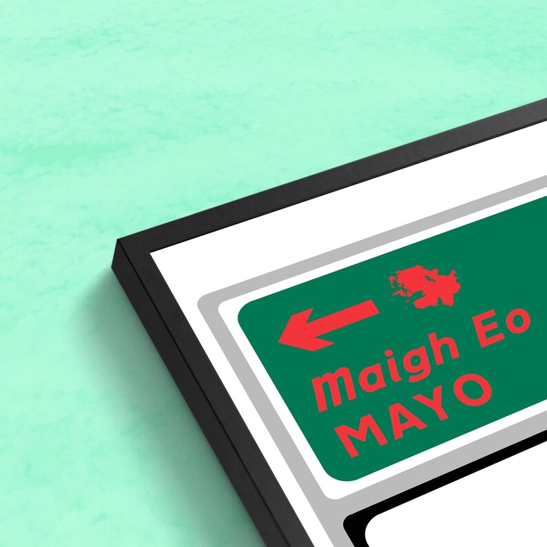 May include: A framed print of a green and white road sign with a red arrow pointing left. The sign reads "maigh Eo MAYO" in red and white text.