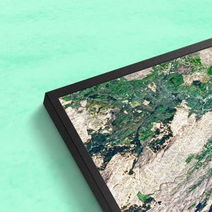 May include: An aerial photograph of a mountainous landscape with a mix of green forests and white rocky terrain. The image is framed in a black picture frame.