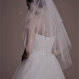 May include: A white bridal veil with a single layer of tulle. The veil is gathered at the top and falls to the shoulders. It is worn by a person with their back to the camera.