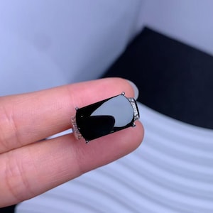 May include: A silver ring featuring a large, rectangular, polished black stone set in the center. The band of the ring is detailed with a textured, rope-like pattern and small, clear, sparkling stones embedded along the sides of the setting.