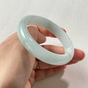 Può includere: Un braccialetto bangle di giada traslucida. Il braccialetto è di colore verde pallido con una superficie liscia e lucida. La giada ha una forma circolare solida e sembra essere una pietra naturale. Il braccialetto è tenuto in una mano.