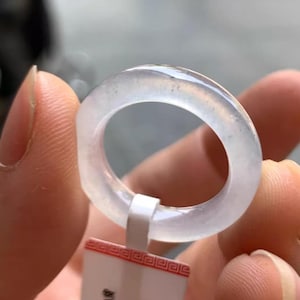 Op de afbeelding: Een doorschijnende, lichtgekleurde ring. De ring wordt omhooggehouden door een wit label met rode accenten. De ring lijkt gemaakt van een glad, gepolijst materiaal. De ring heeft een eenvoudig bandontwerp.