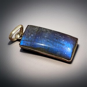 May include: A rectangular pendant featuring a polished labradorite stone with a deep blue iridescent sheen, set in a simple metal frame with a rounded bail attached to the top edge, showcasing natural color variations and a smooth surface.