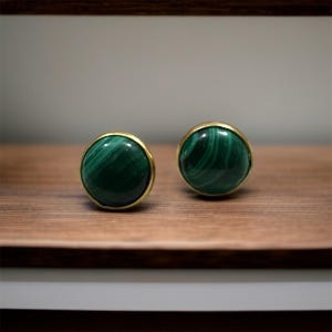 May include: A pair of round stud earrings featuring polished malachite stones with distinct dark and light green swirling patterns, each set within a smooth, thin gold-toned metal bezel frame that encircles the circular gemstone edge.