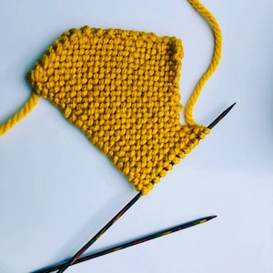 Puede incluir: Una pieza de tejido de punto amarillo con dos agujas de tejer. El tejido está en proceso y la lana todavía está unida.