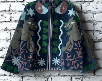 Op de afbeelding: Marineblau geborduurde jas met een hoge kraag, met grillige dieren- en bloemmotieven in groen, roze en bruin. De jas hangt aan een metalen hanger tegen een witte bakstenen muur.