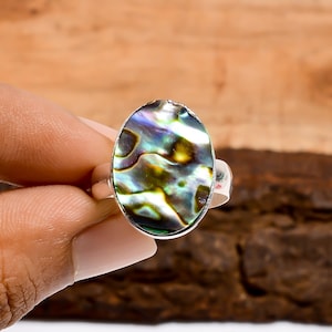 Può includere: Anello ovale in conchiglia di abalone con fascia in argento. Il guscio iridescente presenta vortici di verde, blu e oro. L'anello è tenuto da una mano su uno sfondo di legno. Gioielli in conchiglia di abalone.