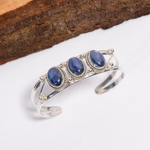 Peut inclure: Bracelet manchette en argent orné de trois pierres bleues ovales. Les pierres sont serties dans un décor argenté avec une bordure tressée. Le bracelet est ouvert et présenté sur une surface blanche.