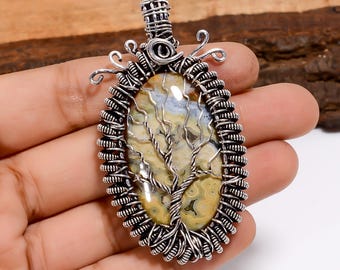 May include: Oval pendant with a silver wire wrap design. The pendant features a tree design over a yellow and cream-colored stone. The pendant is held in a hand.