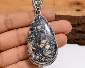 May include: A teardrop-shaped pendant with a silver bail. The pendant features a speckled black, white, and tan stone set in a silver bezel. The stone has a polished, glossy finish. The pendant is held in a hand.