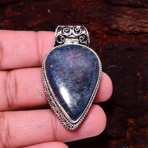 May include: A teardrop-shaped pendant with a blue and purple stone set in a silver-colored metal. The pendant has a decorative bail and a detailed, beaded border. The stone has a polished, reflective surface.
