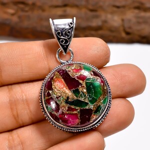Puede incluir: Colgante redondo con un mosaico de piedras rojas, verdes y doradas engastadas en un marco plateado. El colgante tiene una fianza decorativa con un diseño ornamentado. El mosaico de piedras tiene un acabado pulido.
