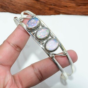 Puede incluir: Una pulsera de plata con tres ópalos ovalados. Los ópalos están engastados en un marco de plata decorativo con un borde en forma de cuerda. La pulsera tiene un diseño sencillo y abierto.