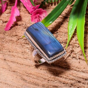 Puede incluir: Un anillo rectangular de piedra preciosa azul con una banda de plata. La piedra preciosa tiene un patrón de rayas y está engastada en un marco de plata con un diseño de cuerda trenzada. El anillo se muestra sobre una superficie de madera.
