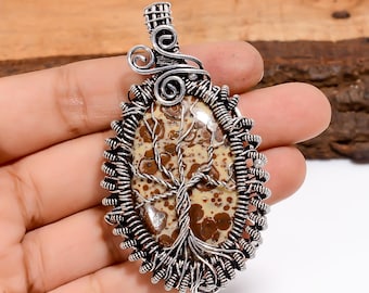 May include: Oval pendant featuring a tree design. The pendant has a brown and beige stone center with a wire tree design. The stone is encased in a silver-colored wire frame. The pendant is held by a silver-colored bail.