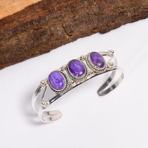 Pode incluir: Uma pulseira de punho prateada com três pedras preciosas ovais roxas. Cada pedra é definida em uma moldura decorativa com contas. A pulseira tem um design aberto, com as extremidades curvadas para fora.