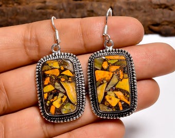 May include: A pair of rectangular earrings with a silver-colored frame and a mosaic-like design. The mosaic features a mix of yellow, orange, and green fragments set against a dark background. The earrings are held in a person's hand.