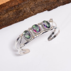 Könnte beinhalten: Silbernes Armreif-Armband mit drei ovalen, irisierenden Edelsteinen. Die Edelsteine zeigen grüne, violette und rosa Farbtöne. Das Armband hat ein detailliertes, offenes Design mit dekorativen Silberakzenten.