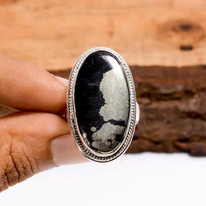 Puede incluir: Anillo ovalado con una piedra negra y plateada. El anillo tiene una banda plateada con un borde texturizado. La piedra tiene una superficie oscura y brillante con motas plateadas. El anillo se muestra sobre un fondo de madera.