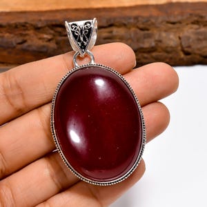 May include: A silver pendant featuring an oval, deep red gemstone. The gemstone is set in a detailed silver frame with a decorative bail. The pendant is held in a hand, showing its size and colour.