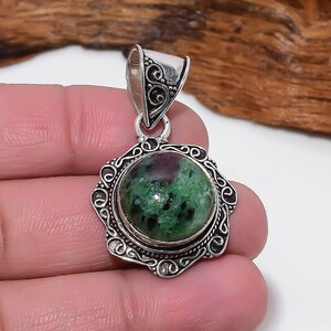 May include: A silver pendant with a round, green and red stone. The stone is set in a decorative, filigree-style frame. The pendant has a bail for attaching to a chain. The pendant is held in a hand.