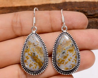 May include: A pair of teardrop-shaped earrings. Each earring features a speckled, light gray and yellow stone set in a silver-colored metal frame with a decorative border. The earrings have a simple hook closure.