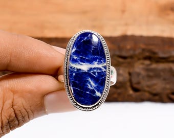 May include: Oval-shaped ring featuring a deep blue stone with white veining, set in a silver-toned metal band. The stone's surface has a polished finish, and the band has a detailed, textured edge. A unique piece of jewelry.