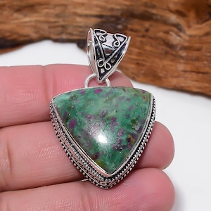 Peut inclure: Un pendentif triangulaire avec une pierre verte et rouge sertie dans un cadre en métal argenté. La pierre a un aspect moucheté et le travail du métal présente des détails complexes. Le pendentif est suspendu à une bélière décorative.