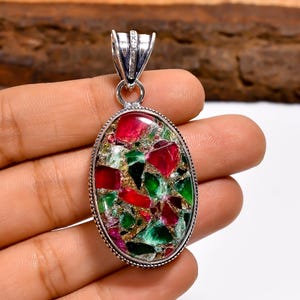May include: Oval pendant with a mosaic of red, green, and gold-toned stones set in a silver-colored metal frame. The pendant has a decorative bail with small, clear stones. The overall design is intricate and colorful, suitable for jewelry.
