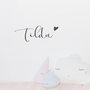 Könnte beinhalten: Eine weiße Wand mit dem Namen "Tilda" in schwarzer Kursivschrift geschrieben, rechts neben dem Namen ein kleines schwarzes Herz. Im unteren Teil des Bildes befinden sich drei Plüschkissen: ein weißes, wolkenförmiges Kissen mit einem lächelnden Gesicht und Wimpern, ein hellrosa, tropfenförmiges Kissen mit einem lächelnden Gesicht und Wimpern und ein hellblaues, tropfenförmiges Kissen mit einem lächelnden Gesicht und Wimpern.