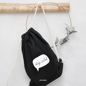 Könnte beinhalten: Schwarze Kordelzugtasche mit weißem Text, der "stay wild!" in einer Sprechblase mit weißen Punkten sagt. Die Tasche hängt an einem Holzhaken mit einem goldenen Metallring.