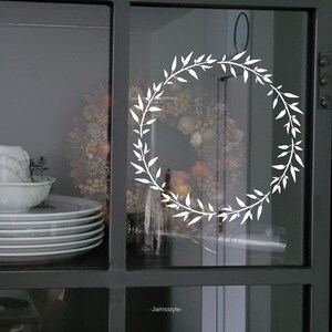 May include: A white wreath decal on a glass cabinet door. The wreath is made of simple leaves and branches.