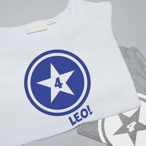 Könnte beinhalten: Weißes T-Shirt mit einem blauen Kreisdesign, das einen weißen Stern mit der Zahl 4 darin zeigt. Der Text "LEO!" ist unter dem Kreis gedruckt.