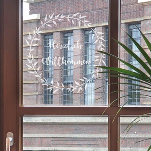May include: A window with a brick building in the background. The window has a white wreath with the words "Herzlich Willkommen" written inside.
