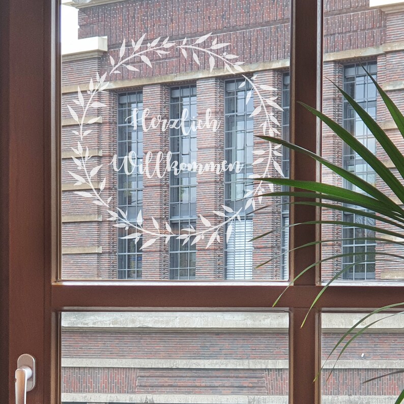 May include: A window with a brick building in the background. The window has a white wreath with the words "Herzlich Willkommen" written inside.