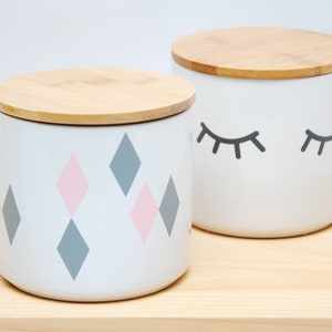 Könnte beinhalten: Zwei Keramik-Aufbewahrungsgläser mit Holzdeckeln. Ein Glas hat ein pink- und graues Diamantenmuster, das andere ein schwarzes Wimpernmuster.