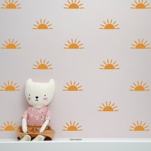 Könnte beinhalten: Ein weißes Stofftier in Form einer Katze sitzt vor einer rosa Wand mit mehreren orangen Sonnenaufklebern. Die Aufkleber sind in einem sich wiederholenden Muster angeordnet.
