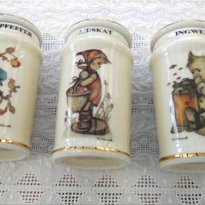 Könnte beinhalten: Drei Vintage-Gewürzgläser aus Keramik mit Goldrand. Jedes Glas zeigt eine andere Illustration von Kindern und ist mit den Namen der Gewürze beschriftet: "Pfeffepfeffer", "Muskat" und "Ingwer".