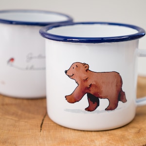 May include: Two white enamel mugs with blue rims. One mug features a brown bear walking on a white background. The other mug has a red bird and the text "Good things take time".