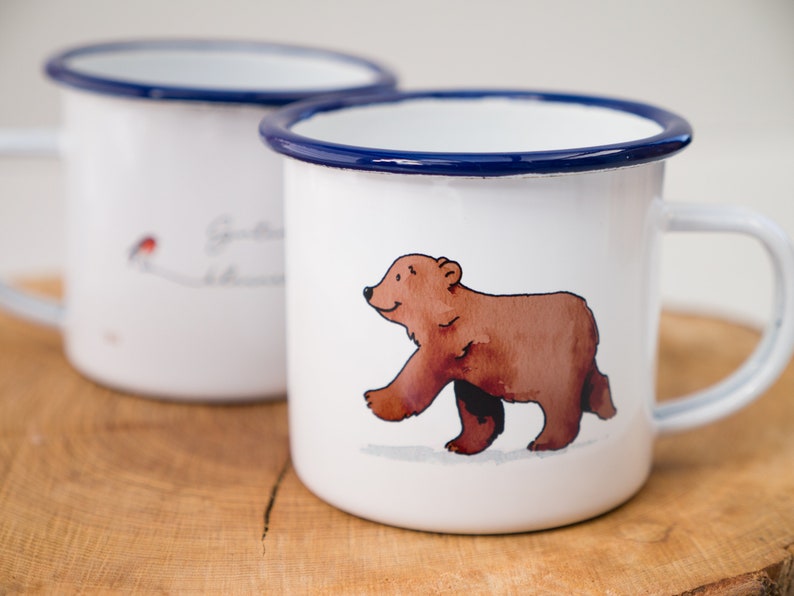 May include: Two white enamel mugs with blue rims. One mug features a brown bear walking on a white background. The other mug has a red bird and the text "Good things take time".