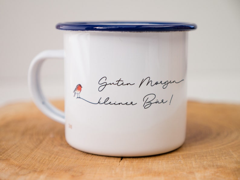 May include: White enamel mug with a blue rim and a robin illustration. The mug has the text "Guten Morgen kleiner B&auml;r!" printed on it.