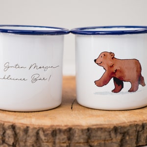 May include: Two white enamel mugs with blue rims. One mug has a red robin and the text "Guten Morgen kleiner B&auml;r!" The other mug has a brown bear walking on a white background.