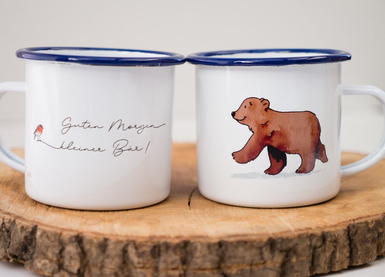 May include: Two white enamel mugs with blue rims. One mug has a red robin and the text "Guten Morgen kleiner B&auml;r!" The other mug has a brown bear walking on a white background.